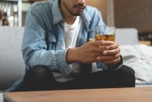 Een man zit thuis op de bank en kijkt somber. In zijn handen heeft hij een glas whisky.